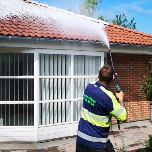 En arbejder fra Glade Fliser iført arbejdsuniform står og sprøjter algebehandlingsmiddel på et rødt tegltag med en højtryksrenser. I baggrunden ses en hvid bygning med et vindue, der har hvide lodrette skodder.